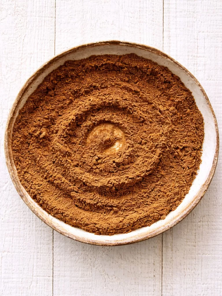 Overhead view of a shallow ceramic bowl filled with a finely ground seven-spice blend, styled on a white timber surface in bright natural light.
