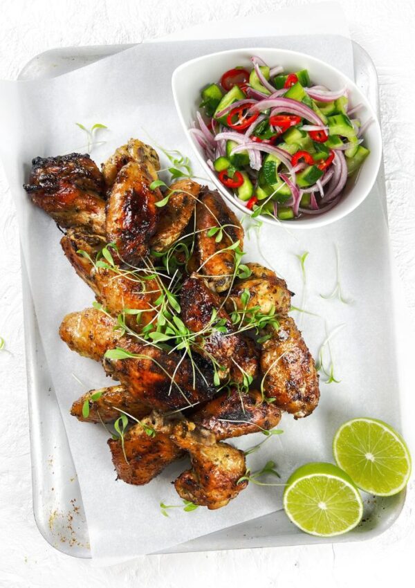 Chicken wings on a tray with lime halves and bowl of cucumber salad