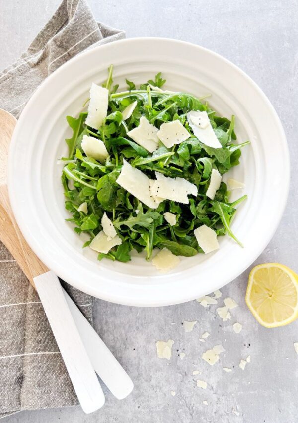 Rocket and parmesan salad in a bowl with lemon half, serving spoons and a tea towel next to it