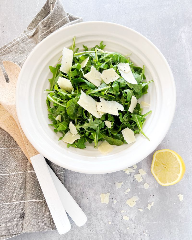 Rocket and parmesan salad in a bowl with lemon half, serving spoons and a tea towel next to it