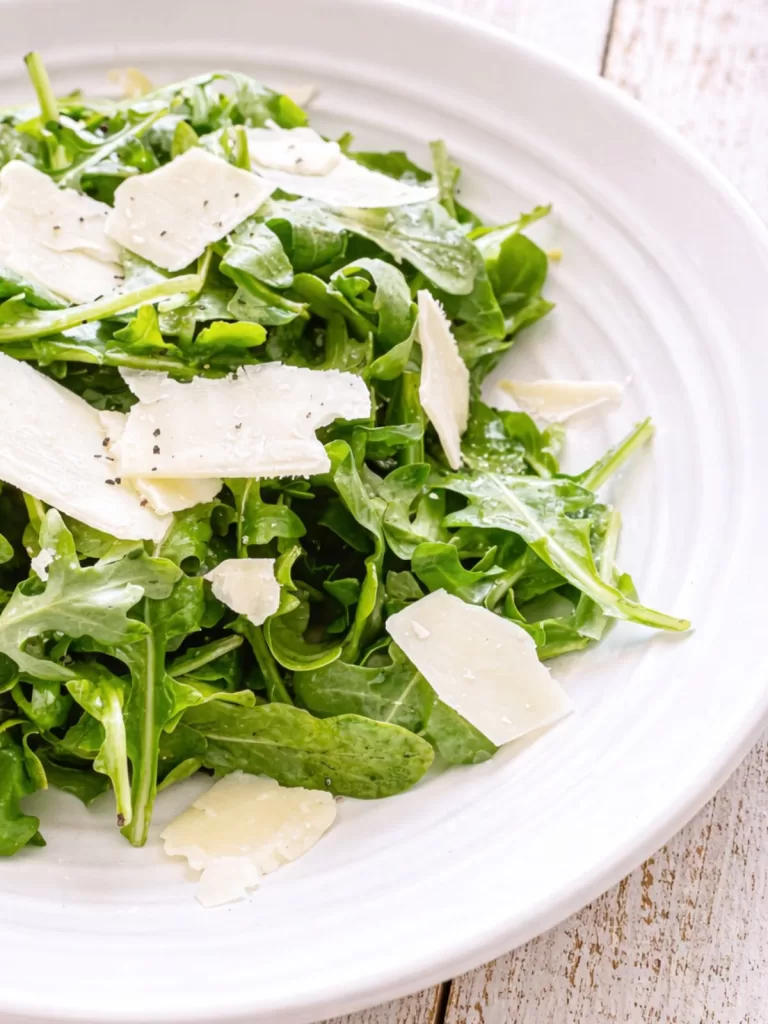Close-up of rocket and shaved Parmesan salad in a white bowl on a rustic white timber background.