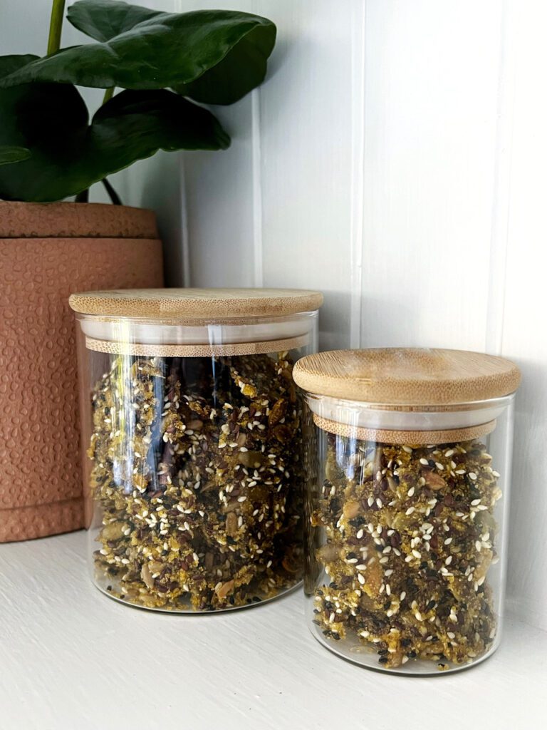 BIRDSEED CRACKERS STORED IN 2 GLASS JARS WITH WOODEN LIDS. PINK POT PLANT IN BACKGROUND WITH GREEN PLANT.