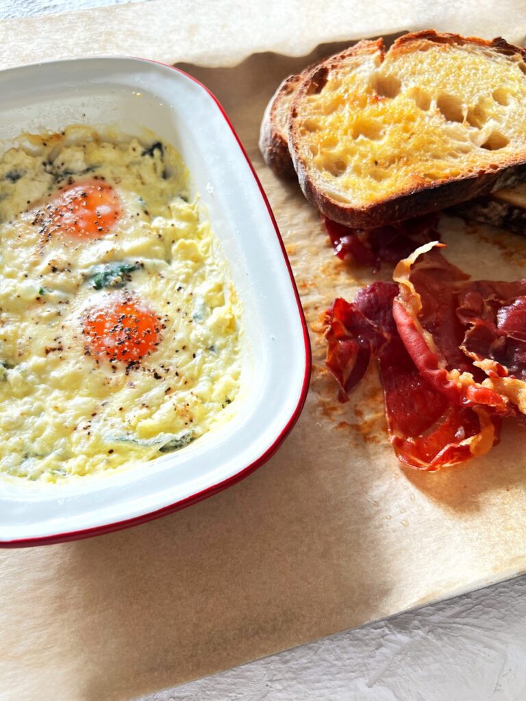 Creamy baked eggs with warrigal greens on baking tray with toast and procsuitto