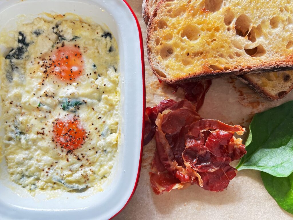 Creamy Baked Eggs in a ramekin with sourdough toast and crispy proscuitto next to it