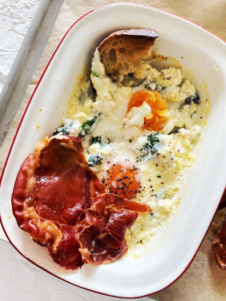 Creamy baked eggs with warrigal greens on baking tray with toast and procsuitto