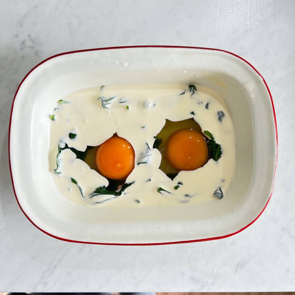 2 eggs in  ramekin with spinach and cream poured around them ready to be baked