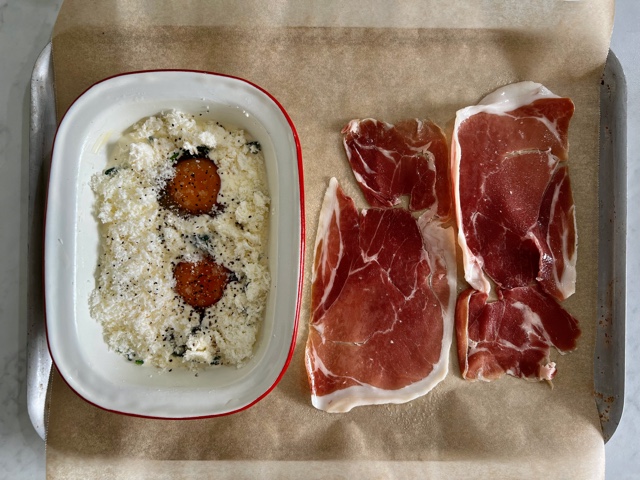 eggs with warrigal greens with cheese on top in a ramekin on a baking tray with raw proscuitto next to it ready to be baked