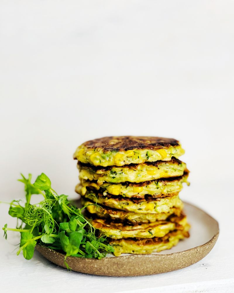 Zucchini & Sweet Corn Fritters stacked on a plate with snow pea sprouts. White background