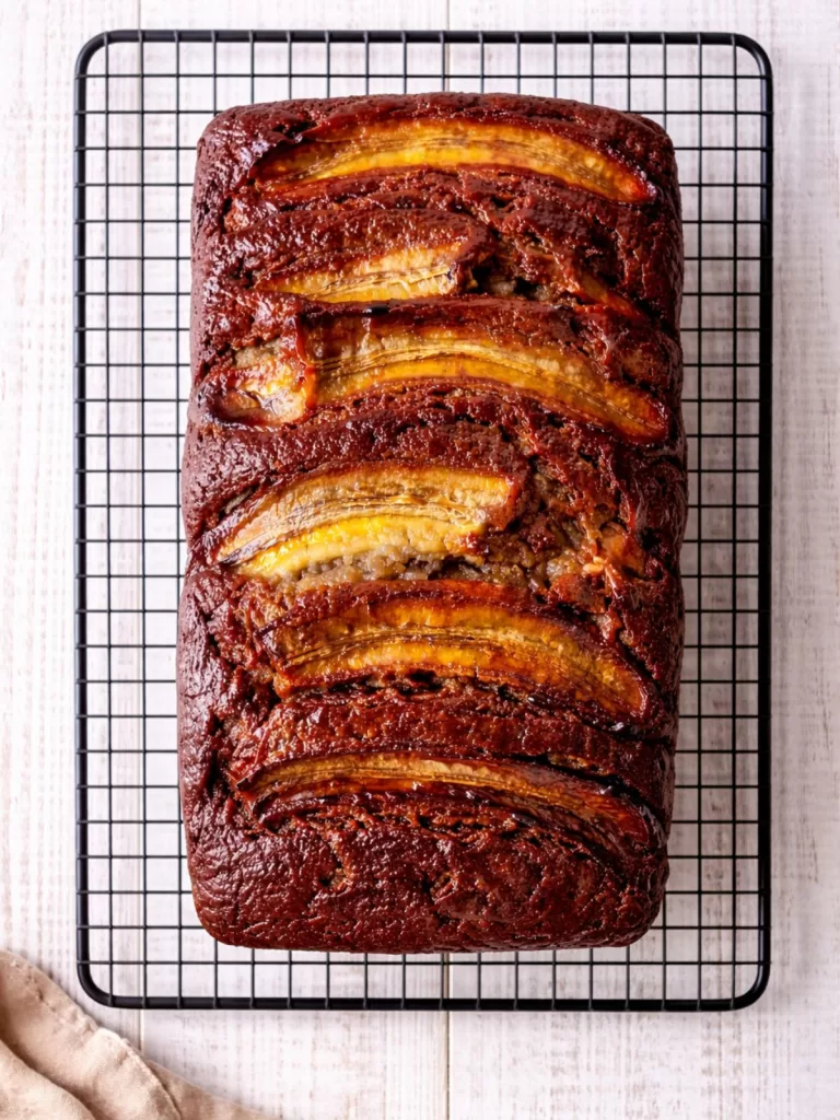 Overhead flat lay of a glossy banana bread loaf with caramelised banana on top, sitting on a cooling rack over a white timber background with a linen cloth in the corner.