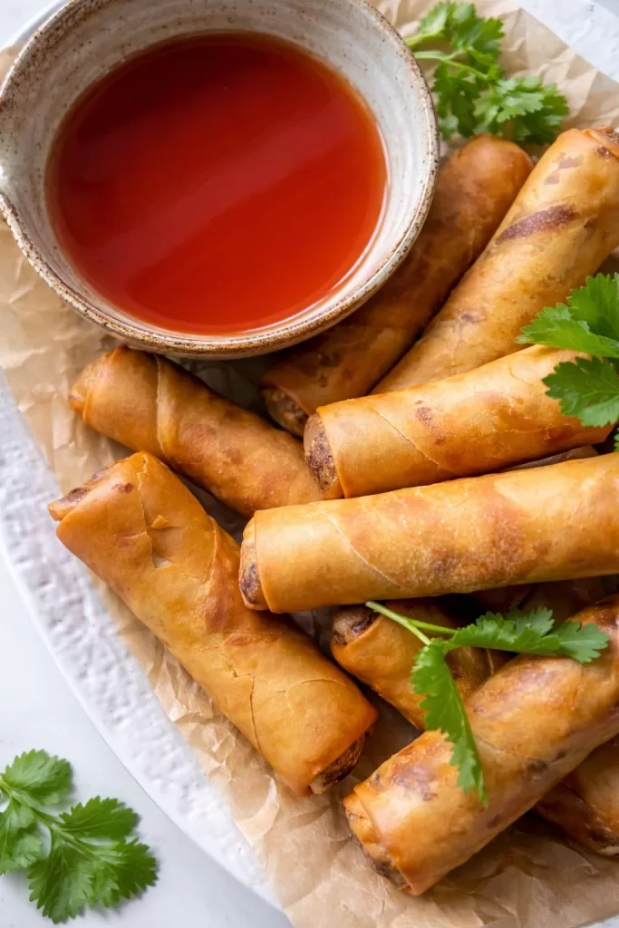 close up of ham and chicken spring rolls on baking paper with a bowl of sweet and sour sauce.