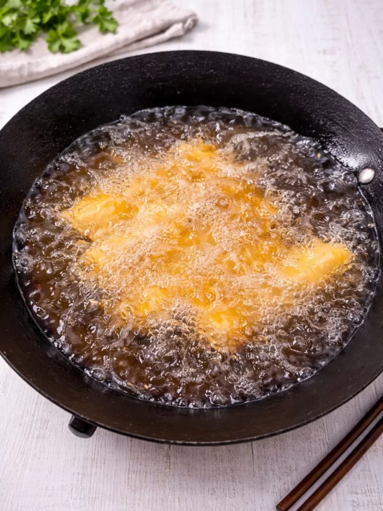 Spring rolls frying in bubbling oil in a black pan on a white timber surface, showing hot oil and crispy golden edges.
