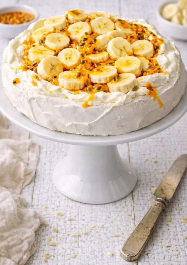 Banana and passionfruit pavlova on a white cake stand, topped with whipped cream, sliced bananas and vibrant orange passionfruit pulp, set on a rustic white timber table with a vintage knife and linen cloth.
