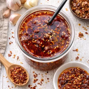 Close-up of a jar of chilli garlic crisp oil filled with red chilli flakes, garlic and sesame seeds in rich red oil, styled on a white timber surface with scattered chilli flakes, garlic cloves, crispy shallots and a small bowl of chilli oil in bright natural light.