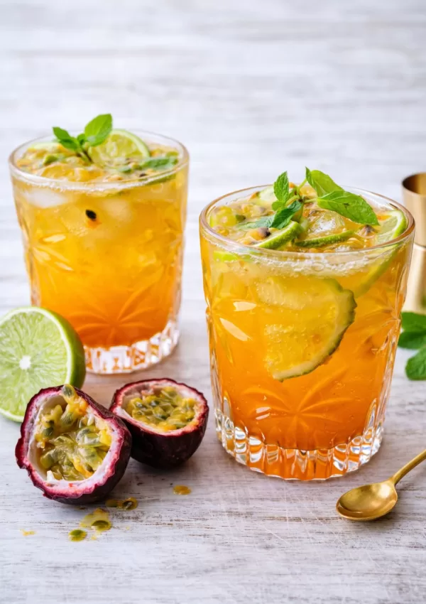 Passionfruit caipiroska with fresh lime and mint served over ice in short glasses on a white timber surface, garnished with halved passionfruit and natural light.