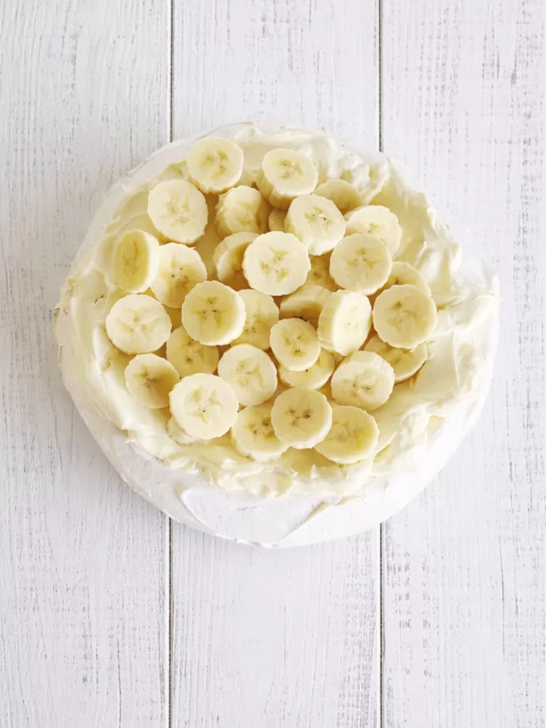 pavlova topped with banana slices on a white rustic timber background