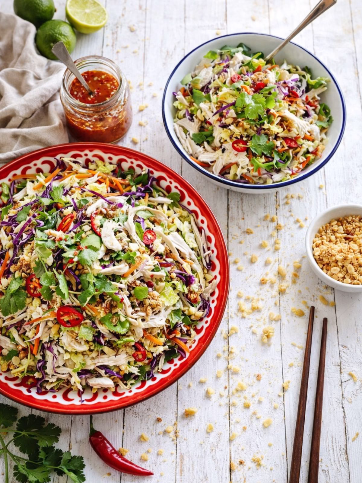 Vietnamese chicken salad served in two bowls on a white timber background, topped with shredded chicken, cabbage, herbs, red chilli and crushed peanuts, with a jar of dressing, fresh limes and chopsticks in natural light.