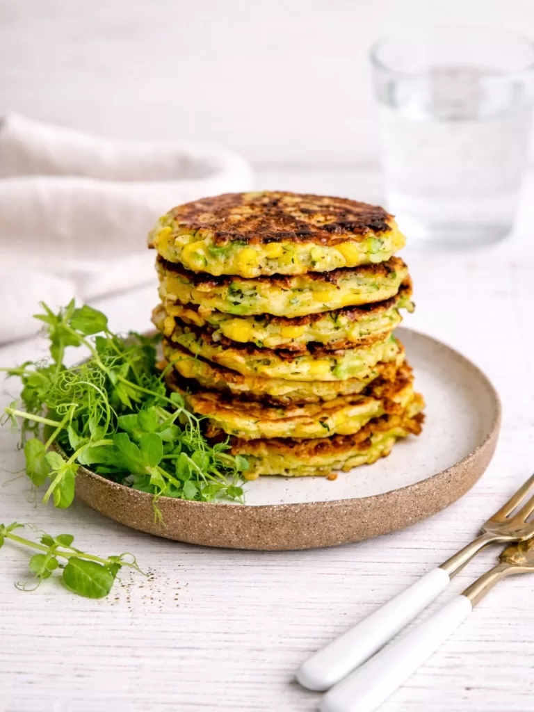 Stack of zucchini and sweet corn fritters on a ceramic plate with fresh greens, a napkin and a glass of water on a white timber background.