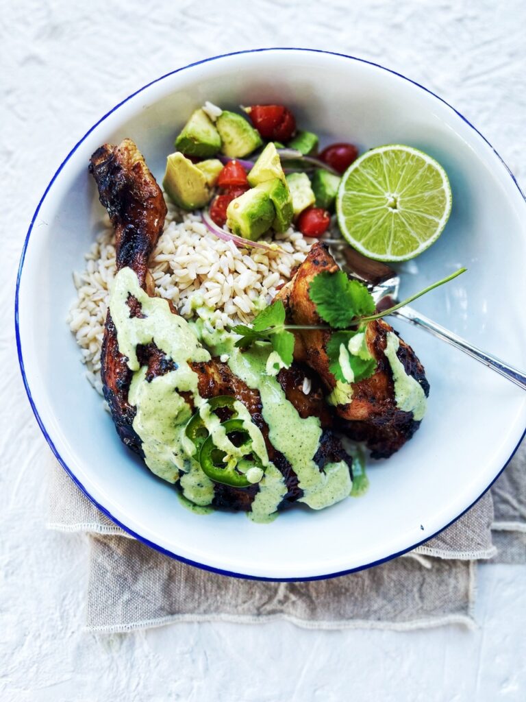 Peruvian Roast Chicken pieces in a bowl with green sauce drizzled over it. Lime wedge, rice and avocado salsa .