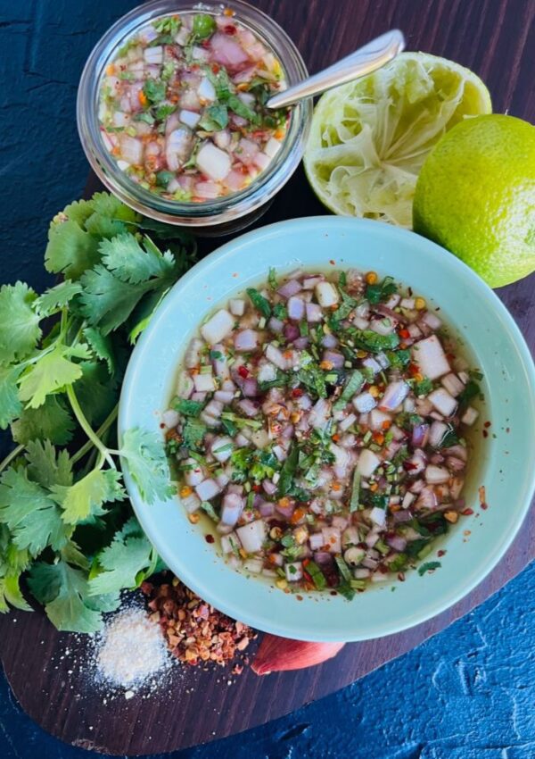 Nam Jim Jaew Dipping Sauce in a bowl. Lime wedges, coriander, chilli and shallots on display around the bowl of sauce