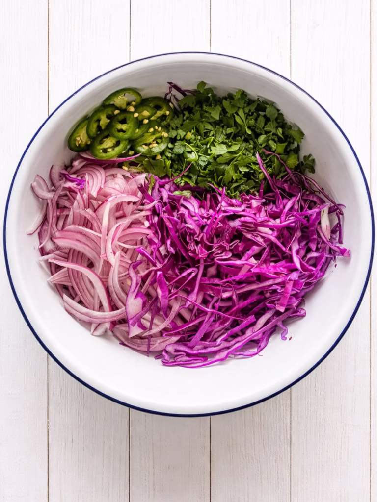 Overhead view of a white enamel bowl with red cabbage, sliced red onion, jalapeno, and chopped parsley on a white timber surface.