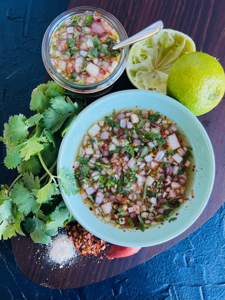 Nam Jim Jaew Sauce in bowl and also in a glass jar. Ingredients used are displayed around the outside. Lime, Coriander, Shallot, Chilli and ground rice powder