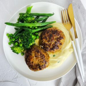 Beef & Vegetable Rissoles with mashed potato and steamed green beans, broccoli and peas on a white plate with white and gold cutlery on white textured background