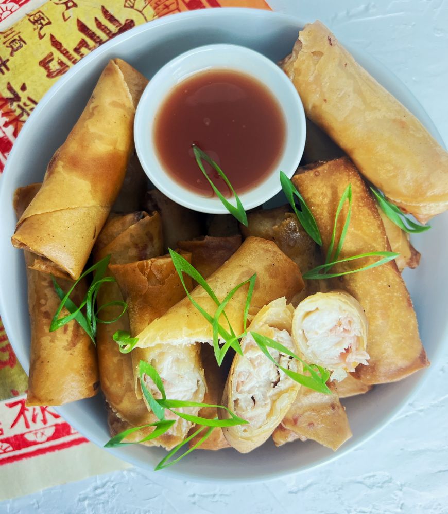 CHICKEN SPRING ROLLS IN WHITE BOWL WITH SWEET AND SOUR SAUCE. GARNISHED WITH SPRING ONIONS