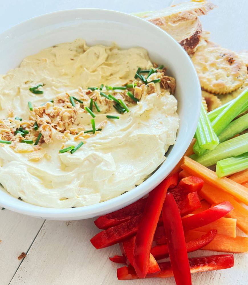 Curried egg dip in a white bowl sprinkled with crispy onions and chives. Crackers and vegetable sticks behind it