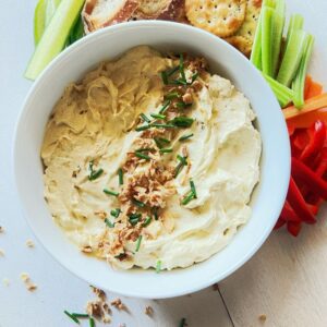 Curried Egg Dip in a white bowl with vegetable sticks behind it. The dip is sprinkled with fried onions and chives.