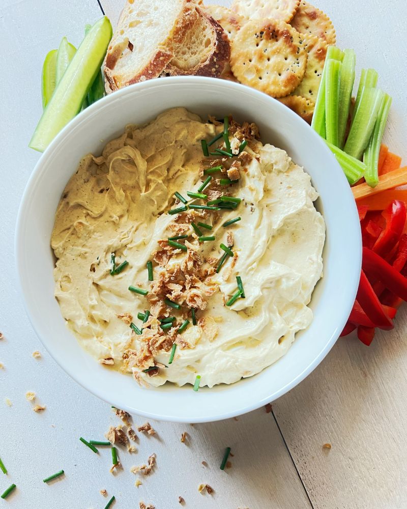 curried egg dip in white bowl with vegetable sticks and crackers behind it. 