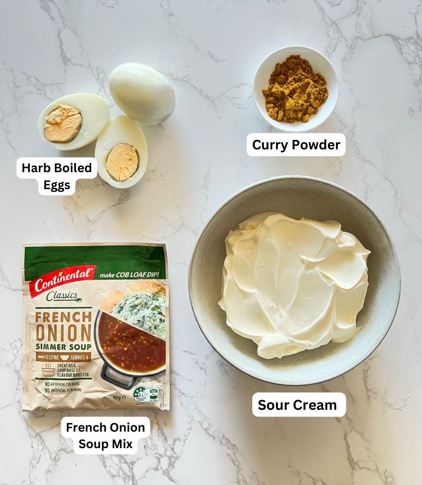 Ingredients board for egg dip: hard boiled eggs, curry powder, french onion soup mix and sour cream laid out on a marble board
