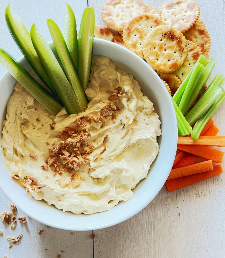 Dip in a white bowl with cucumber sticks sticking out of it. Crackers and vegetable sticks in the background.