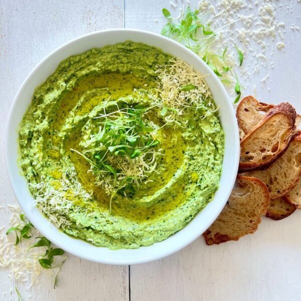 Fresh Spinach Dip in a white bowl garnished with parmesan, olive oil and microgreens. Bread slices next to bowl on white wood background
