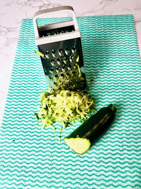 green kitchen cloth with a box grater, some grated zuccchini and 1/2 a zucchini sitting on it