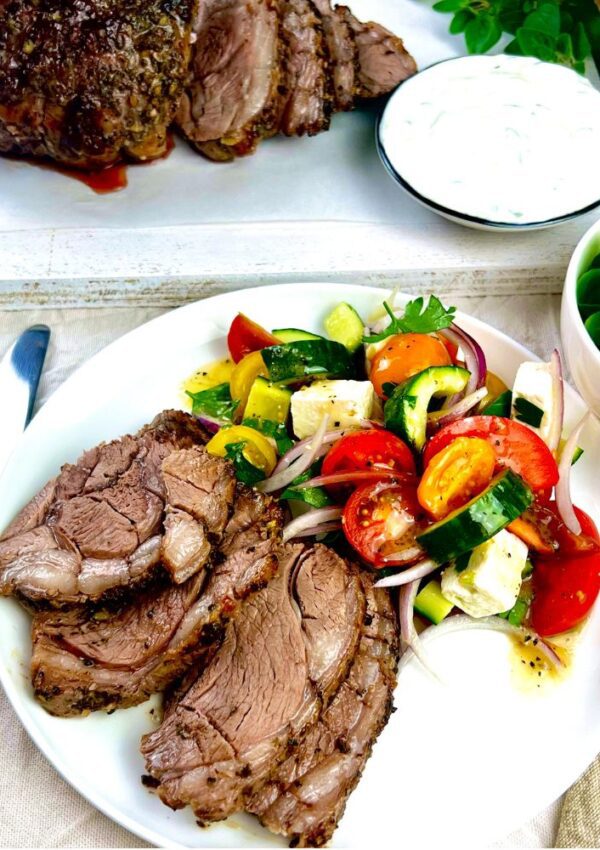 white dinner plate with sliced lamb and greek salad. Bowl of yoghurt and lamb on board in background