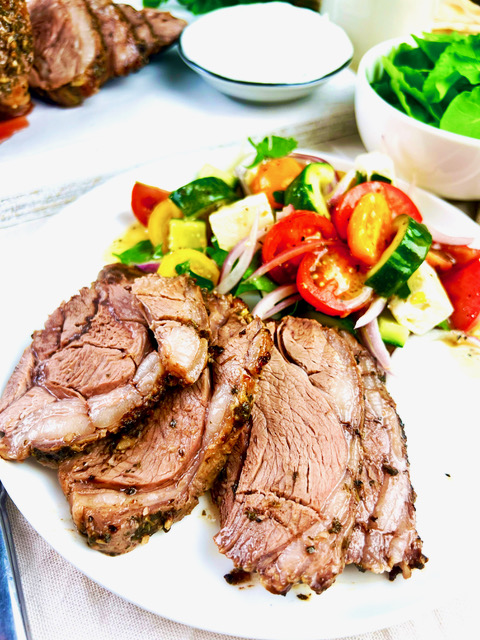 Greek sliced lamb leg on white plate with Greek salad. small bowl of yoghurt and rocket leaves in background