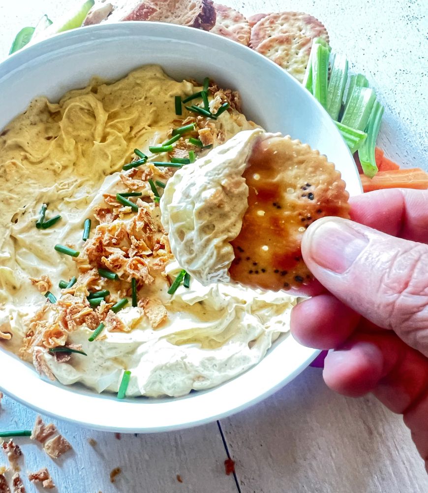 Person scooping up dip on a biscuit with bowl of dip in the background.