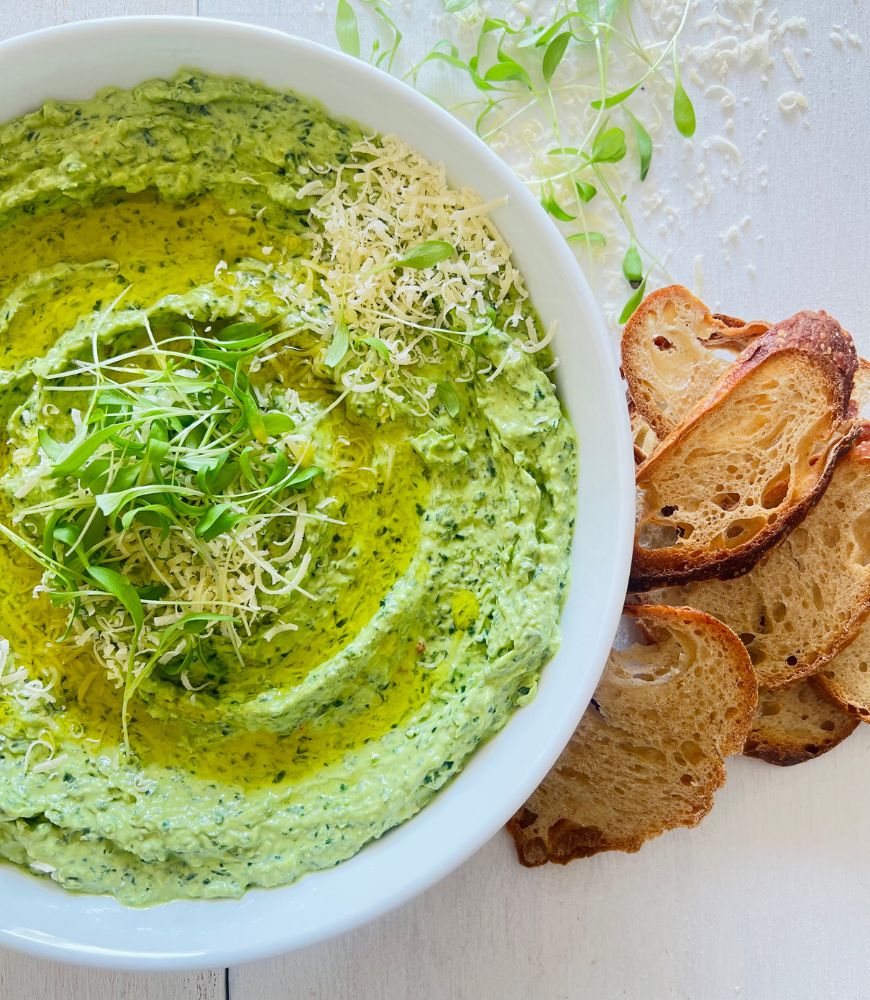 creamy fresh spinach dip in a white bowl with crispy thin bread slices to the side. Grated parmesan and microgreens scattered on top and sides