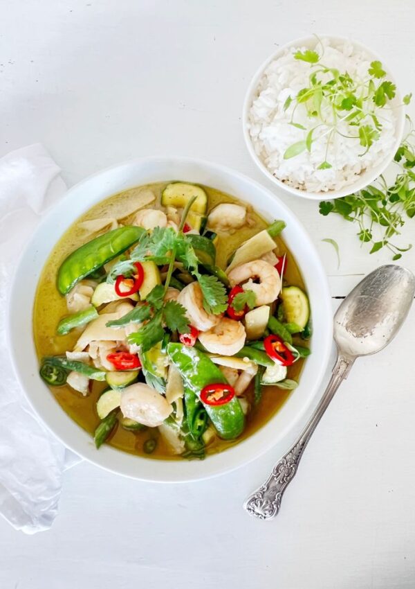 THAI GREEN PRAWN CURRY IN A WHITE BOWL ON WHITE BACKGROUND WITH SMALL BOWL OF RICE AND A SILVER SPOON NEXT TO IT