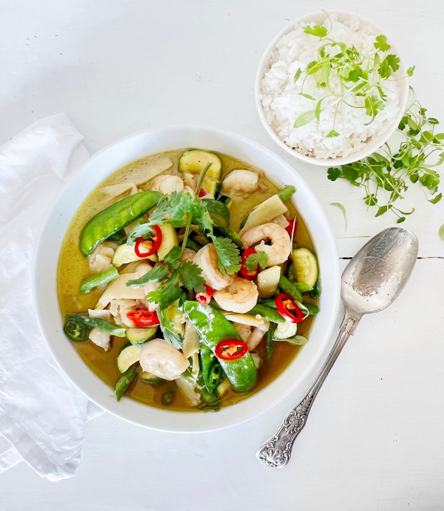 Thai Green Prawn Curry in white bowl with white napkin under it, a sliver spoon to the right and a small bowl of rice garnished with micro-greens