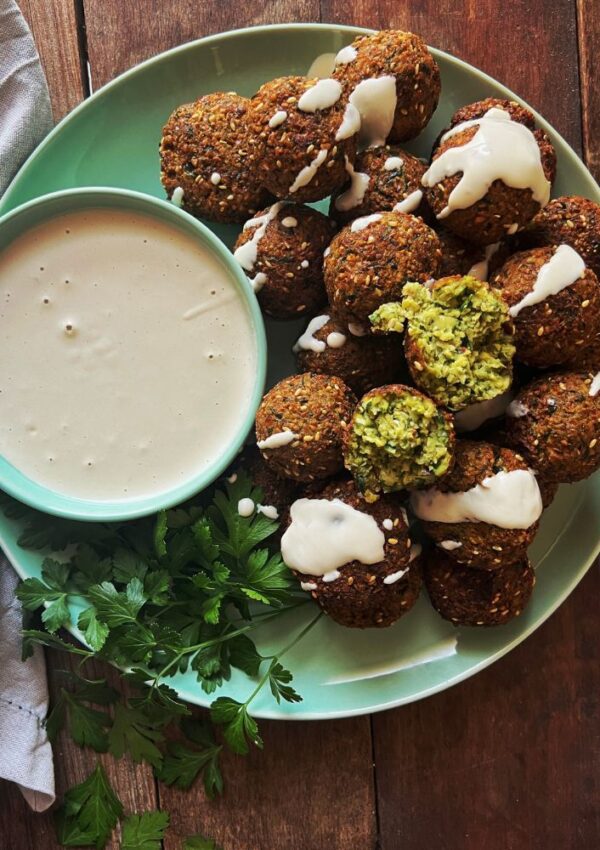 Zucchini & Sesame Falafels on a green plate with a bowl of tahini dipping sauce and some parsely as garnish. Set on a wooden background