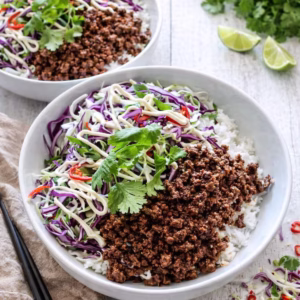 Asian beef bowls with rice, seasoned mince, and cabbage slaw in white bowls on a white timber background with fresh herbs and lime in natural light