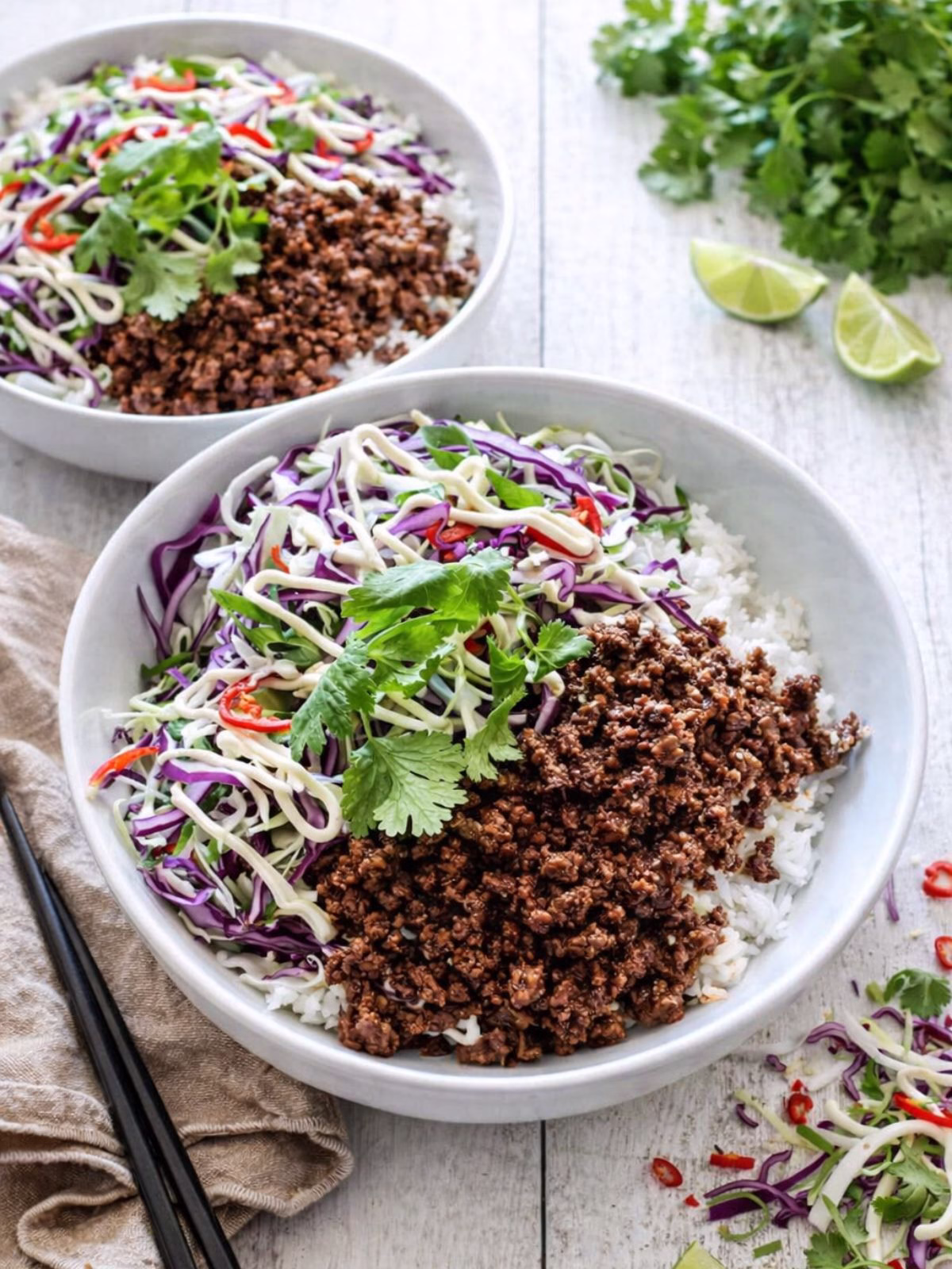 Asian beef bowls with rice, seasoned mince, and cabbage slaw in white bowls on a white timber background with fresh herbs and lime in natural light