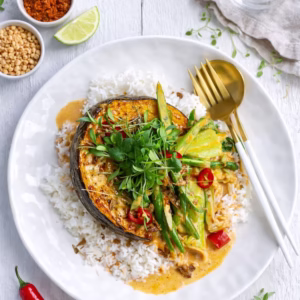 Overhead view of baked pumpkin Thai red curry served with jasmine rice, green beans, bok choy, sliced red chilli, fresh herbs, and creamy curry sauce on a white plate with gold cutlery on a white timber table.