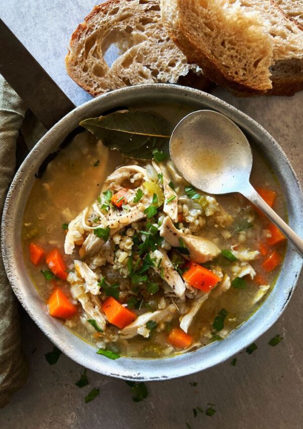 Chicken, Barley & Freekeh Soup in small pan with bread and soup spoon and green cloth to side. Grey background