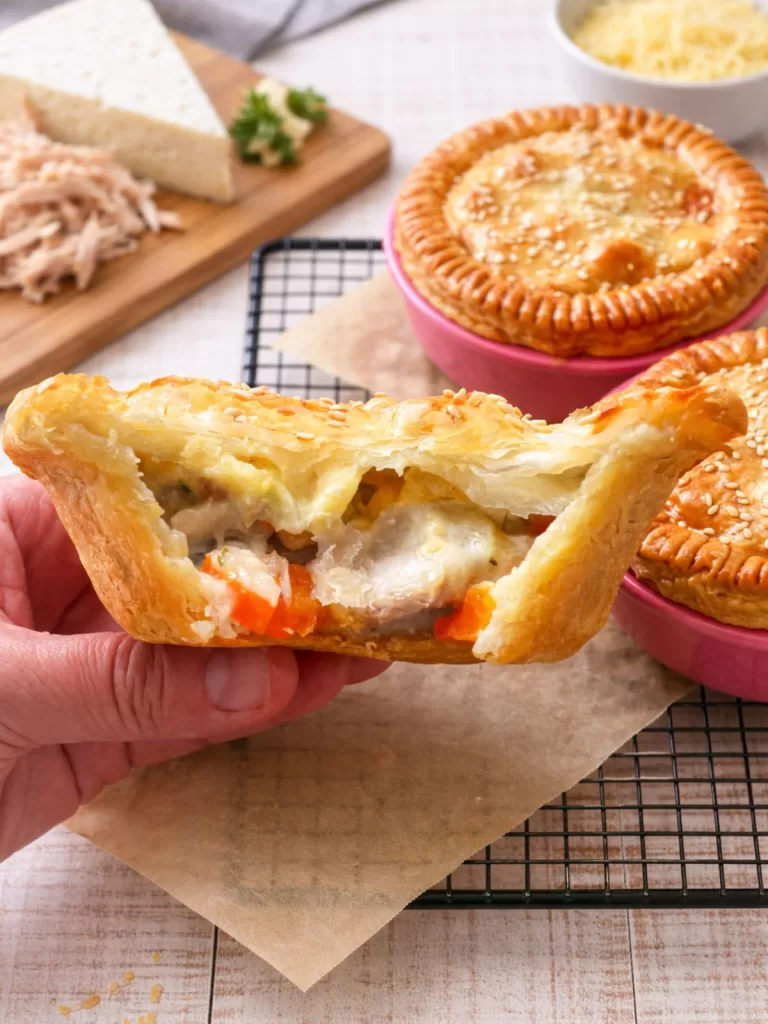 Hand holding a halved chicken and Camembert pie with creamy chicken, carrot and mushroom filling visible inside a golden sesame-topped pastry, with whole pies and cheese in the background on a white timber surface.