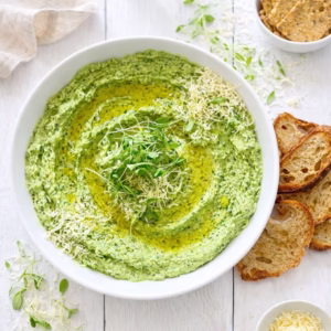 Creamy fresh spinach dip in a white bowl topped with olive oil, parmesan, and microgreens served with toasted sourdough on a white timber table