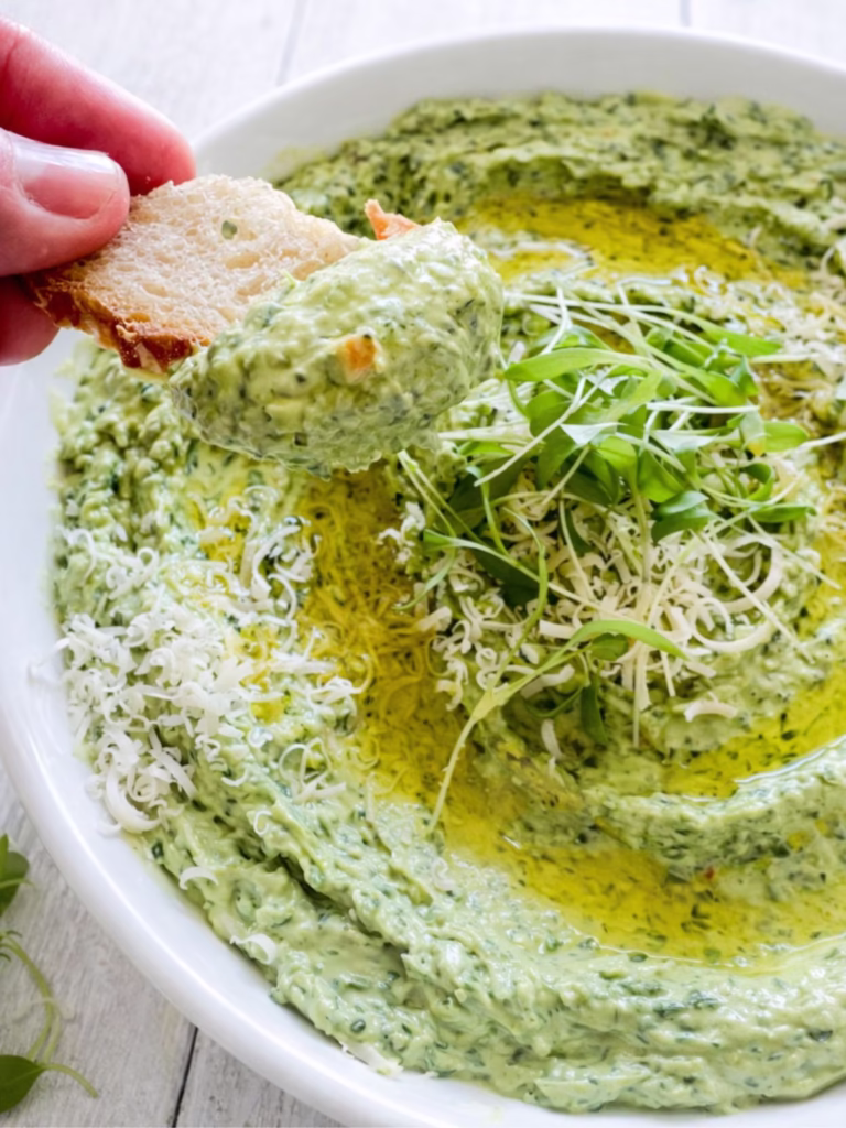 Close up of creamy fresh spinach dip being scooped with sourdough bread, topped with olive oil, parmesan, and microgreens on a white timber table