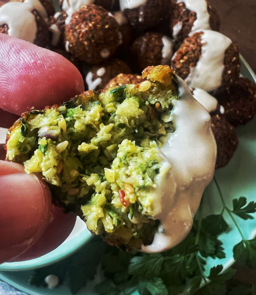 fingers holding a zucchini falafel with tahini dip on it