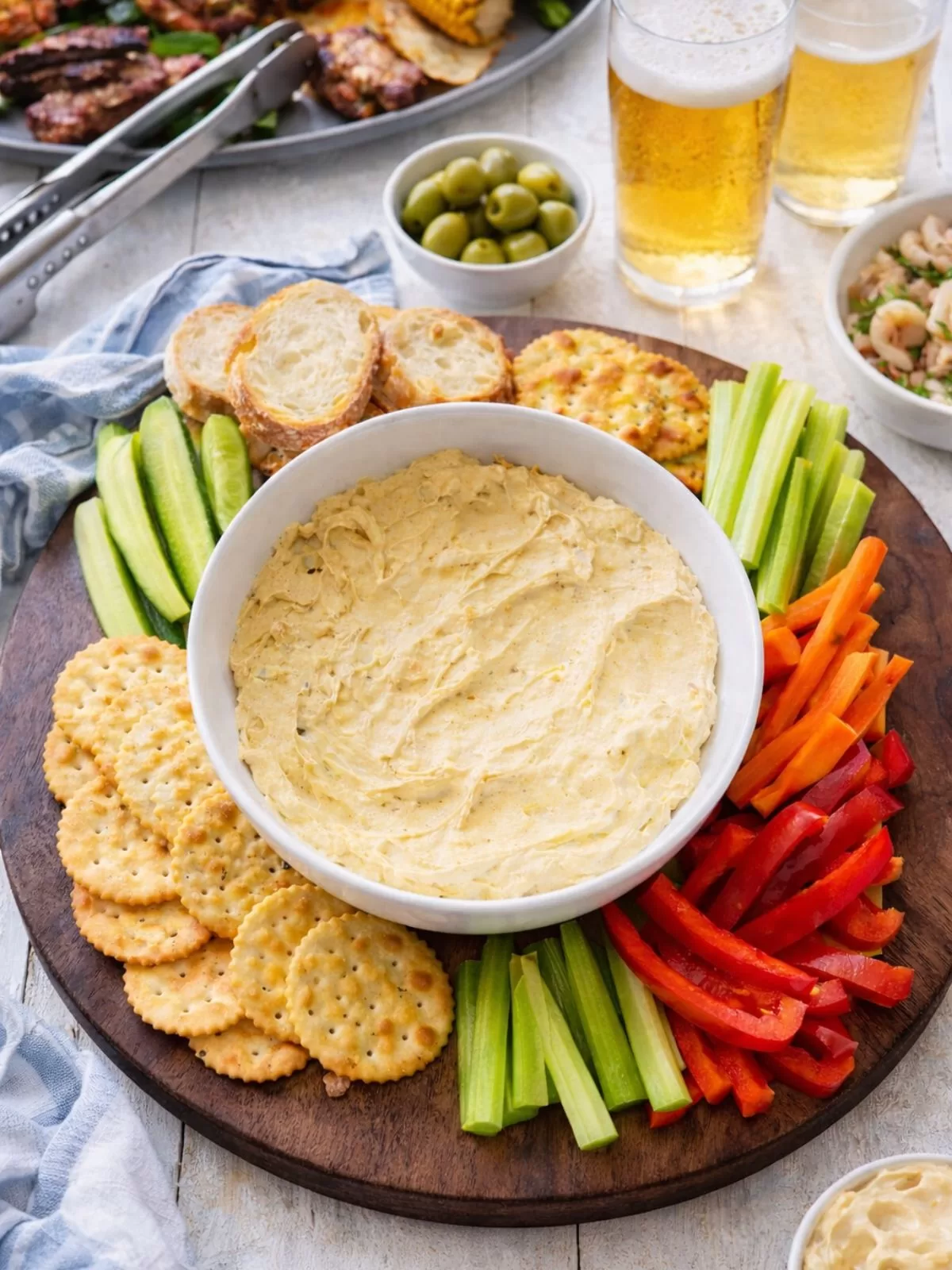 Curried egg dip in a white bowl on a round dark wooden board with crackers, bread slices and fresh vegetable sticks, topped with chunky chopped egg and a light sprinkle of curry powder, set on a white timber table with BBQ dishes and drinks in the background.